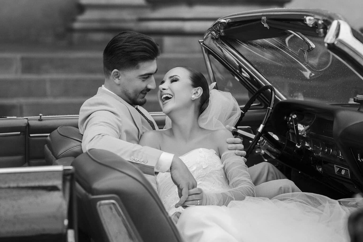Sort-hvidt billede af et brudepar i en vintage cabriolet; bruden ler, brudgommen ser på hende