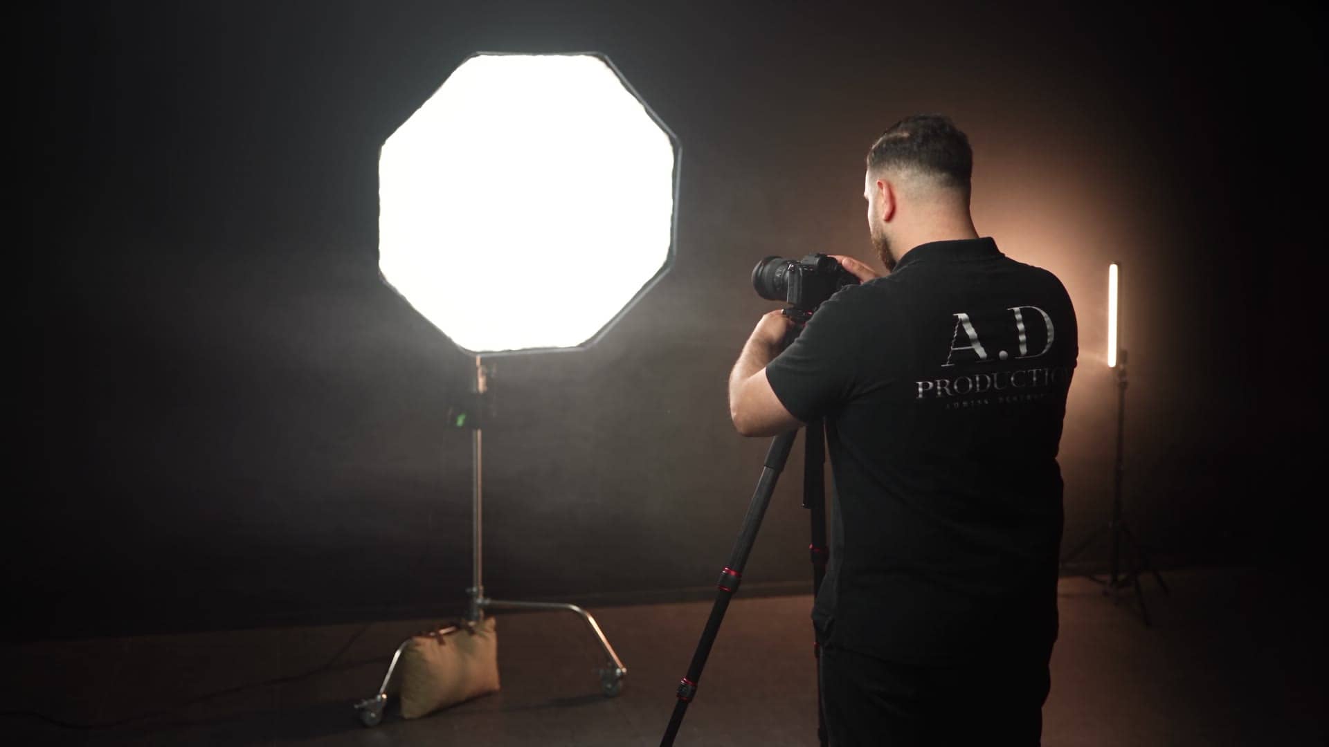 Fotograf set bagfra ved kamera på stativ, mens en stor softbox lyser varmt i et mørkt studie